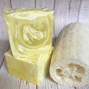 May include: Three bars of yellow and white swirled soap are stacked next to a natural loofah sponge. The soap has a marbled design. The loofah is a light beige color and has a cylindrical shape.