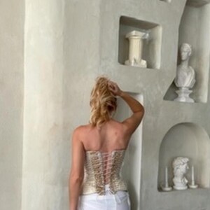 May include: A strapless corset top featuring a textured, metallic gold and cream woven fabric. The back of the garment is secured with a vertical lace-up closure using thin, light-colored cord, creating a fitted silhouette against the torso.