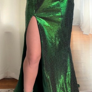 May include: A floor-length, emerald green sequin gown with a high slit. The dress has a fitted silhouette that flares out slightly at the bottom. The fabric shimmers, reflecting light. The dress is suitable for formal events.