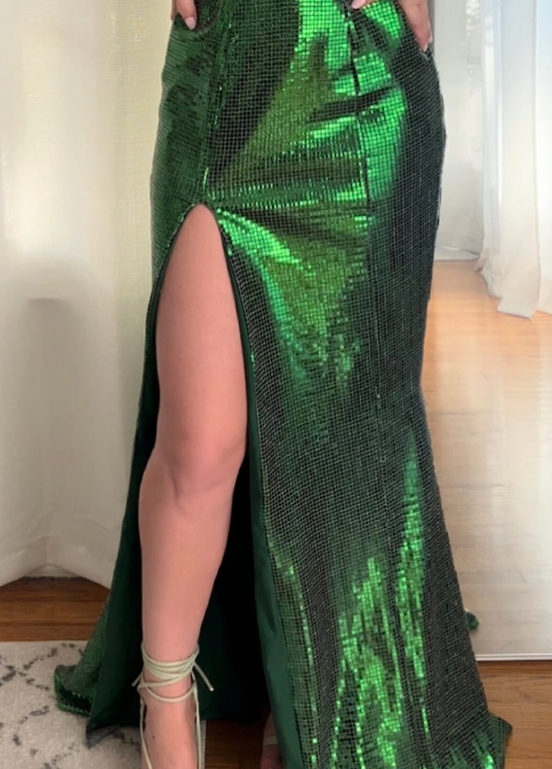 May include: A floor-length, emerald green sequin gown with a high slit. The dress has a fitted silhouette that flares out slightly at the bottom. The fabric shimmers, reflecting light. The dress is suitable for formal events.