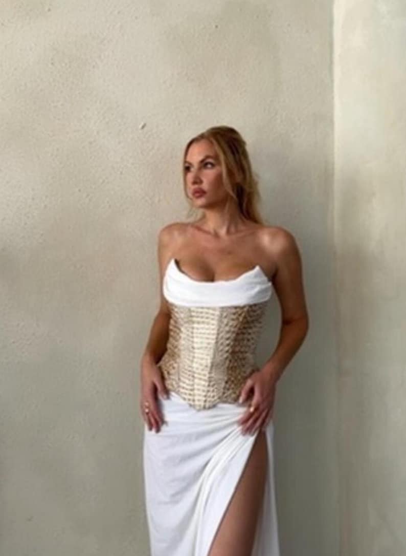 May include: A strapless corset top featuring a white folded neckline and a gold textured bodice with a structured, boned design, paired with a long white skirt that includes a high side slit, creating a two-piece ensemble with a fitted silhouette.
