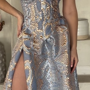 May include: A strapless, light blue and gold snakeskin-patterned dress with a fitted bodice and a flowing skirt. The skirt has a high slit on the left side.