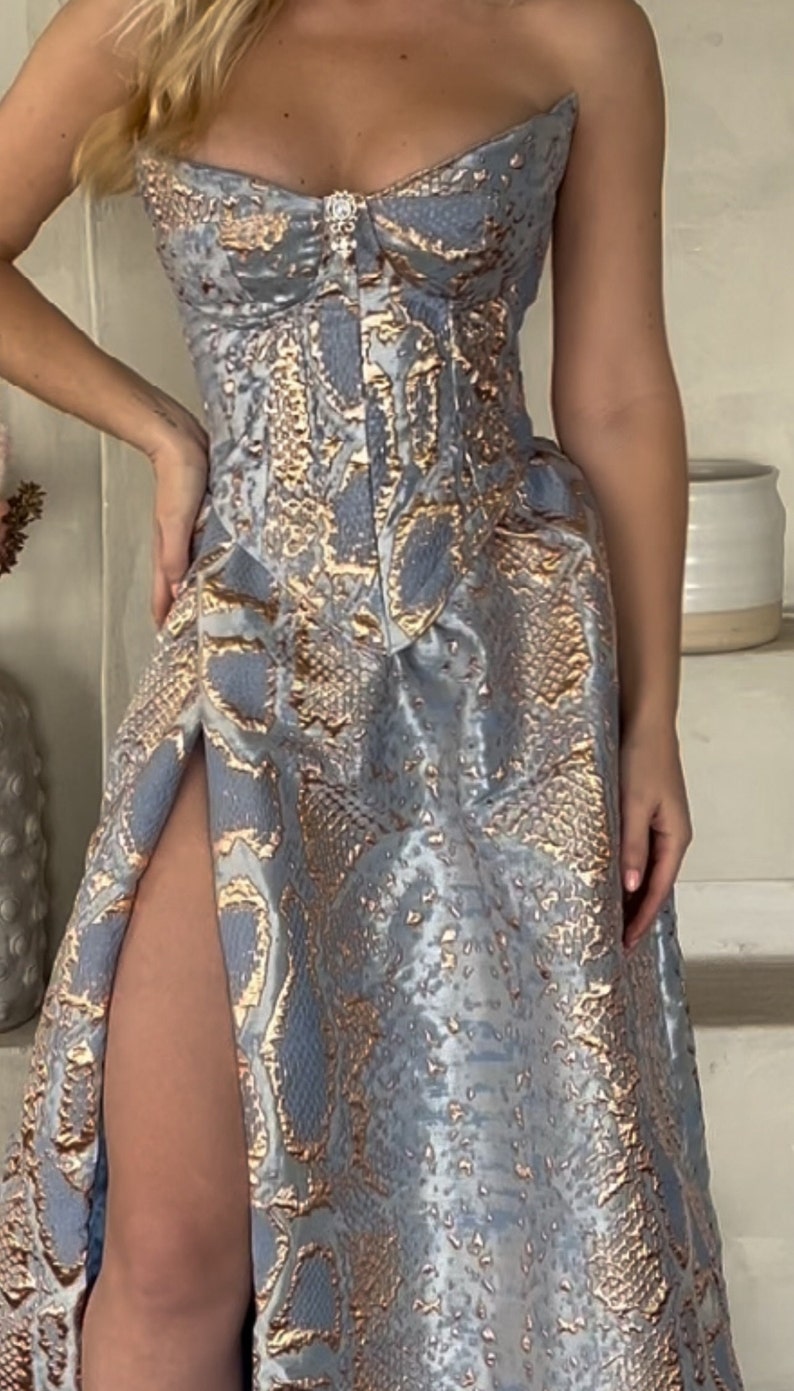May include: A strapless, light blue and gold snakeskin-patterned dress with a fitted bodice and a flowing skirt. The skirt has a high slit on the left side.