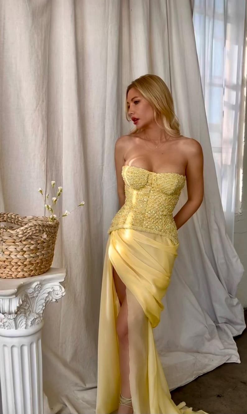 May include: A woman wearing a yellow strapless dress with a beaded bodice and a flowing skirt. The dress has a high slit on the side. The woman is standing in front of a white curtain.