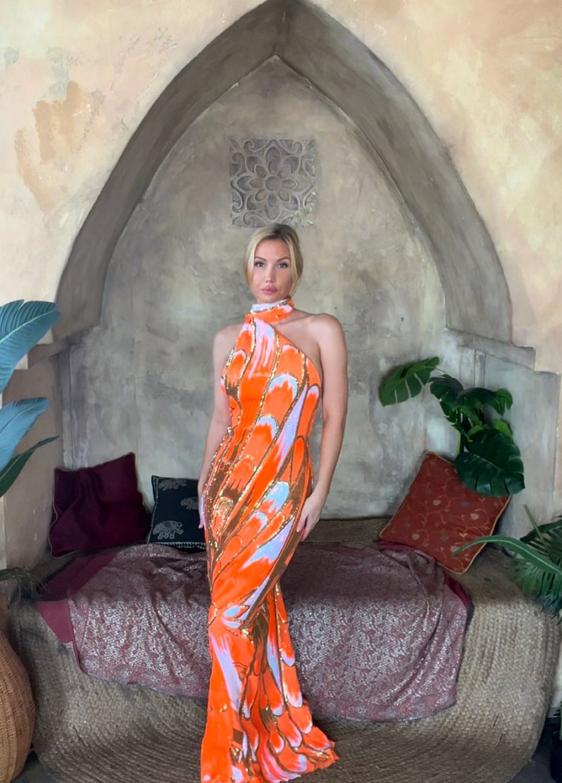 May include: A woman wearing a long, orange and white halter dress with a sequined pattern. The dress has a high neckline and a fitted silhouette. The woman is standing in front of a stone archway with a decorative tile.