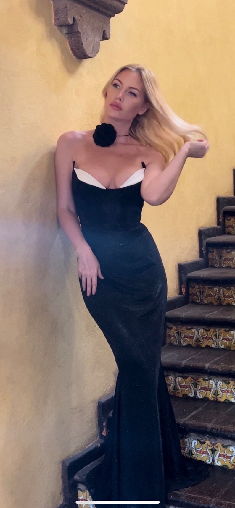May include: A woman wearing a black strapless gown with a white trim and a black flower on her neck. She is standing on a staircase with patterned tiles.