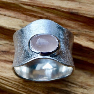 May include: A silver ring with a light pink oval gemstone set in the center. The ring has a textured, hammered finish.