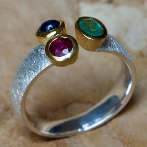 Könnte beinhalten: Ein silberner Ring mit strukturierter Oberfläche, besetzt mit drei Edelsteinen in Gold. Die Edelsteine sind ein blauer Saphir, ein roter Rubin und ein grüner Smaragd.