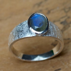 Könnte beinhalten: Ein silberner Ring mit einem blauen, ovalen Edelstein, der in einem strukturierten Band gefasst ist.