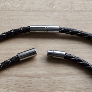 Puede incluir: Un brazalete de cuero negro con cierre magnético plateado. El brazalete está hecho de cuero trenzado y tiene un diseño sencillo.