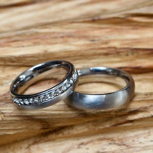 Puede incluir: Dos alianzas de boda plateadas, una con una fila de pequeños diamantes, sobre una superficie de madera.