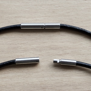 Op de afbeelding: Een zwarte leren koordketting met zilveren magnetische sluitingen. De ketting is open en de sluitingen zijn gescheiden.