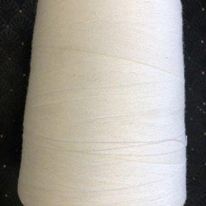 May include: A large spool of white yarn, tightly wound and conical in shape. The yarn appears to be a soft, textured material, suitable for knitting or crocheting projects. The spool is set against a dark background.