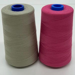 May include: Two spools of thread. One is a muted green color, and the other is a vibrant magenta. Both spools have a blue plastic core at the top. The thread is tightly wound around the spools, creating a conical shape. These are likely for sewing or crafting.