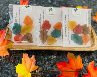 May include: Three clear cellophane bags filled with colorful, leaf-shaped soaps. The soaps are in shades of orange, yellow, red, brown, and green. The bags are on a wooden tray, with fall leaves around them. Text on the bags reads "Specializing in Gift Boxes".