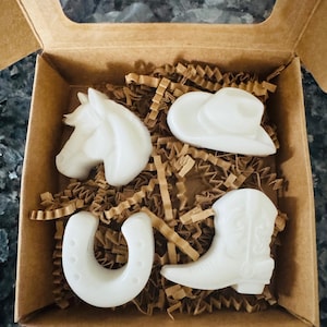 May include: A set of four white cowboy-themed soap bars in a brown cardboard box. The shapes include a horse head, cowboy hat, horseshoe, and cowboy boot, nestled on brown crinkle paper.