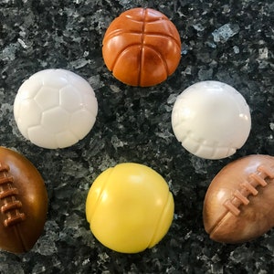 May include: Six different sports balls made of chocolate. The balls are a basketball, soccer ball, baseball, tennis ball, and two footballs. The balls are all different colors and have the appropriate markings for each sport.