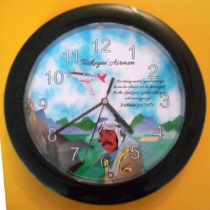 May include: A black framed clock with a colorful design featuring a plane, a pilot, and a sky background. The clock face has Roman numerals and the text "Tuskegee Airmen" and a Bible verse from Joshua 1:9 NIV.
