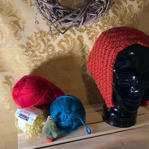 May include: A red crocheted hat with ear flaps, displayed on a black mannequin head.  The hat is sitting on a wooden shelf with two balls of yarn, one red and one blue, and a small artificial Christmas tree.