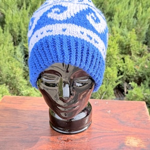 May include: A knit beanie featuring a blue and white wave pattern across the crown and a solid blue ribbed brim. The hat is displayed on a black mannequin head, showcasing the contrasting color scheme and the detailed texture of the knit fabric.