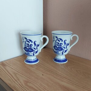 Peut inclure: Deux tasses en céramique blanches avec des motifs floraux bleus. Les tasses ont une base en piédestal et une anse. Le motif bleu représente un panier de fleurs et de feuillage. Les tasses sont sur une surface en bois.