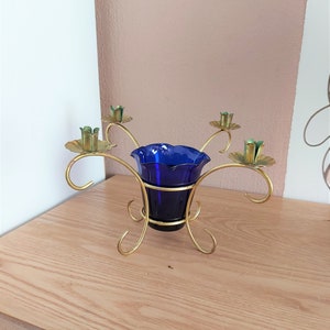 May include: A vintage candle holder featuring a cobalt blue glass cup. The gold-coloured metal frame has four curved arms, each holding a small candle cup with a green accent. The candle holder sits on a wooden surface. The dimensions are approximately 15 cm wide.