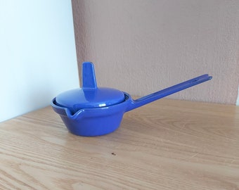 May include: A blue ceramic gravy boat with a lid and a handle. The boat has a spout for pouring.