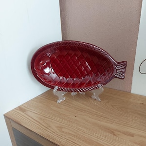 Può includere: Un piatto da portata in ceramica rossa a forma di pesce con un design in rilievo testurizzato. Il piatto è su un supporto in plastica trasparente.