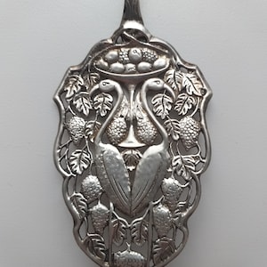 Puede incluir: Utensilio para servir plateado con un diseño ornamentado. El utensilio presenta una forma de escudo decorativo con dos pájaros, uvas y hojas. Un cuenco de fruta está en la parte superior. El mango es largo y delgado.