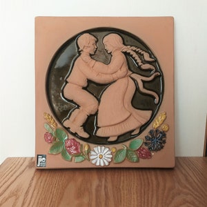 Puede incluir: Un azulejo de terracota con un diseño en relieve de una pareja bailando. La pareja está en un círculo rodeado de flores y hojas. El azulejo está firmado "JIC Sweden".