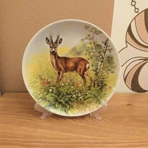 May include: A decorative ceramic plate with a detailed illustration of a deer in a natural setting. The plate shows a brown deer in a field of green grass and flowers, with trees in the background. The plate is displayed on a clear plastic stand.