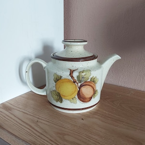 Puede incluir: Una tetera de cerámica blanca con tapa y asa marrones. La tetera tiene un esmalte moteado y un diseño de frutas marrones y amarillas.