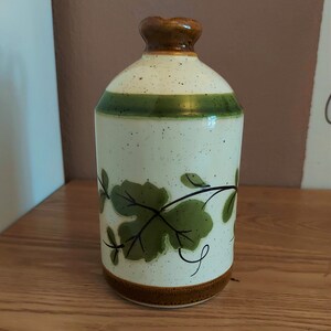 May include: A ceramic bottle with a speckled off-white body, a green band, and a brown top and base. The bottle features green leaf and vine designs. The bottle is sitting on a wooden surface.