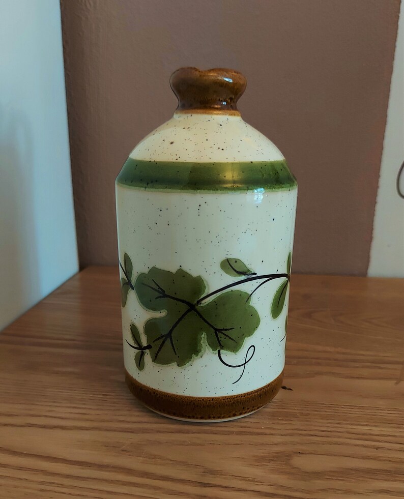 May include: A ceramic bottle with a speckled off-white body, a green band, and a brown top and base. The bottle features green leaf and vine designs. The bottle is sitting on a wooden surface.