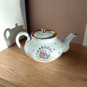 Könnte beinhalten: Eine weiße Teekanne mit goldenem Deckel und Verzierungen. Die Teekanne hat ein florales Design mit einem Paar in der Mitte. Die Teekanne steht auf einer Holzoberfläche.