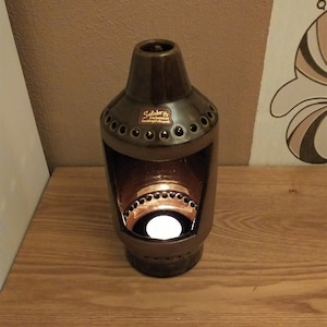 Puede incluir: Un portavelas de cerámica marrón con forma cilíndrica y una amplia abertura. Presenta una serie de pequeños agujeros en la parte superior y los lados. Una vela encendida en el interior emite un brillo cálido. El texto "Salobo" es visible.