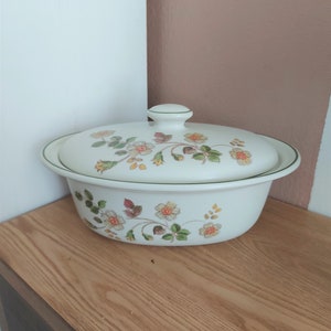 以下が含まれることがあります： ピンク、黄色、緑色の花柄で装飾された、蓋付きの白い楕円形のキャセロール皿。縁とハンドルは緑色のトリムです。木製の表面に置かれています。料理の提供に適しています。