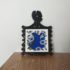 Puede incluir: Salvamanteles de hierro fundido negro con un diseño de gallo en la parte superior y una baldosa de cerámica blanca con una jarra azul y flores blancas en el centro.