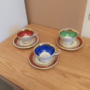 Könnte beinhalten: Drei Teetassen mit goldenen Verzierungen und unterschiedlichfarbigen Innenräumen: rot, blau und grün. Jede Tasse steht auf einer passenden Untertasse mit einem goldenen und weißen Blumenmuster.