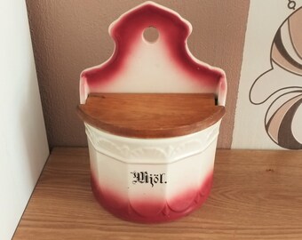 Puede incluir: Un tarro de especias de cerámica blanca para colgar en la pared con un diseño degradado en rojo y blanco. El tarro tiene una tapa de madera y la palabra "Wjör." impresa en la parte delantera.
