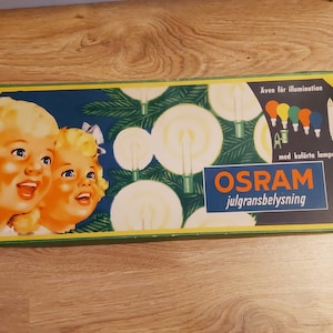 Op de afbeelding: Een vintage doos met OSRAM kerstverlichting met een feestelijk ontwerp met twee kinderen en een kerstboom. De doos heeft de tekst "Även för illumination med kulörta lampor" en "OSRAM julgransbelysning".