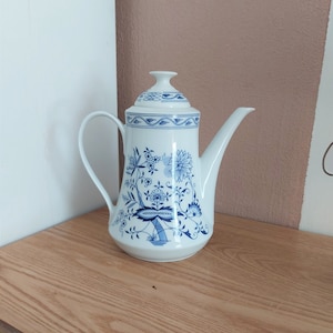 May include: White ceramic coffee pot with a blue floral design. The pot has a long spout, a curved handle, and a lid. The floral pattern includes flowers, leaves, and other botanical elements. The pot is sitting on a wooden surface.