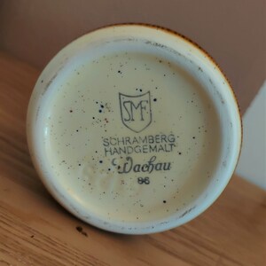 May include: A ceramic mug base with a speckled cream interior and a brown rim. The base features the text "SCHRAMBERG HANDGEMALT Wachau 86" and a shield logo.