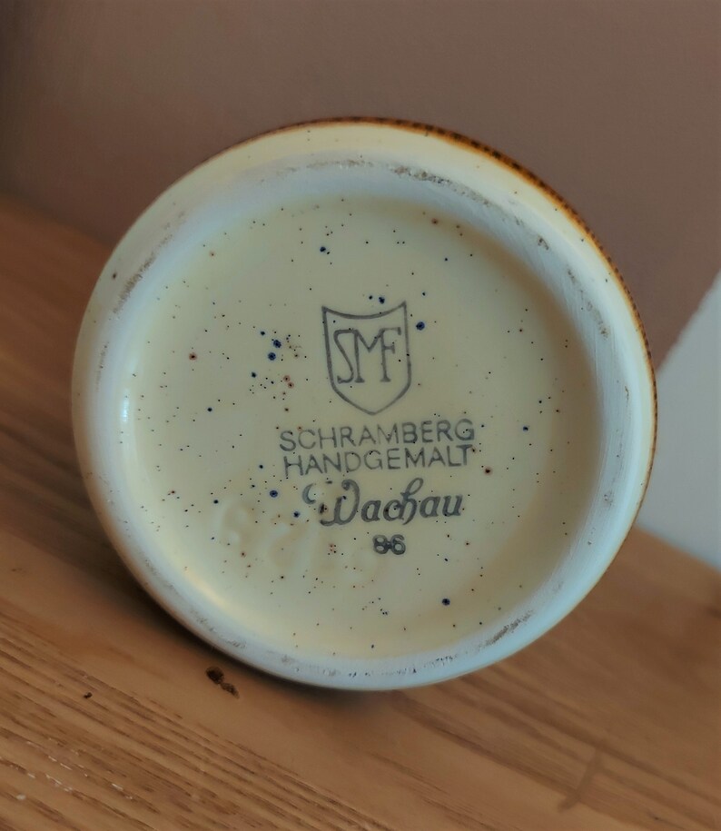 May include: A ceramic mug base with a speckled cream interior and a brown rim. The base features the text "SCHRAMBERG HANDGEMALT Wachau 86" and a shield logo.