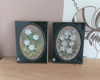 May include: Two ceramic wall plaques with white flower designs. Each plaque has a dark green border with a beaded edge, framing an oval relief of flowers and leaves. The background is a tan colour. The plaques are rectangular.