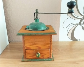 Puede incluir: Un molinillo de café vintage de madera con una tapa de metal verde y un cajón. El molinillo tiene una manivela y una perilla de madera en el cajón.