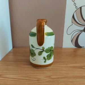 May include: A ceramic jug with a brown handle and base, and green accents. The jug features a white body with green leaf and vine designs. The jug is sitting on a wooden surface.