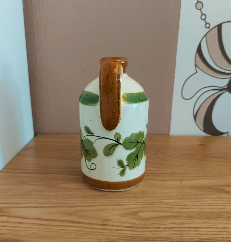 May include: A ceramic jug with a brown handle and base, and green accents. The jug features a white body with green leaf and vine designs. The jug is sitting on a wooden surface.