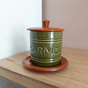 Può includere: Vaso per marmellata in ceramica verde con coperchio e base in legno. La parola "MARMEL" è in rilievo sulla parte anteriore. Il coperchio e la base sono di colore marrone caldo. Il vaso è posto su una superficie di legno.