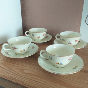 Könnte beinhalten: Vier cremefarbene Tassen und Untertassen mit Blumendesigns. Jede Tasse und Untertasse hat einen Goldrand. Das Blumenmuster umfasst rote, blaue und gelbe Blumen. Das Set ist auf einer Holzoberfläche angeordnet.